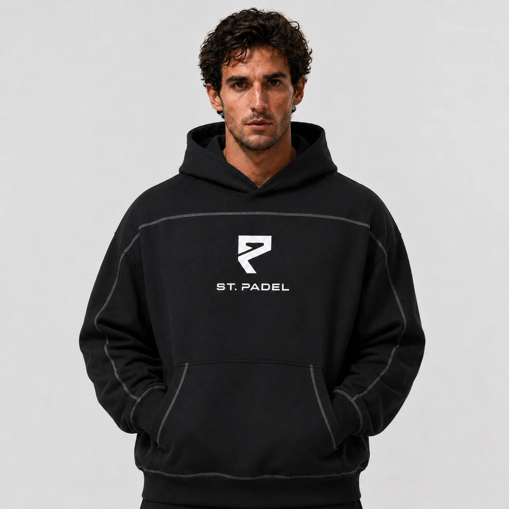 St. Padel Boxy Stich Hoodie. A man wearing a black pullover hoodie with a white 'ST. PADEL' logo and stylized 'P' emblem on the chest, photographed against a neutral light gray background.