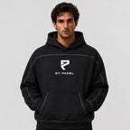 St. Padel Boxy Stich Hoodie. A man wearing a black pullover hoodie with a white 'ST. PADEL' logo and stylized 'P' emblem on the chest, photographed against a neutral light gray background.