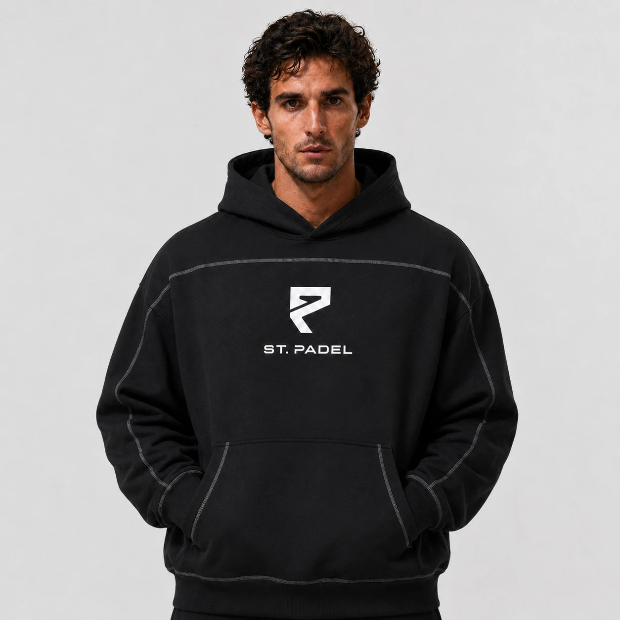 St. Padel Boxy Stich Hoodie. A man wearing a black pullover hoodie with a white 'ST. PADEL' logo and stylized 'P' emblem on the chest, photographed against a neutral light gray background.