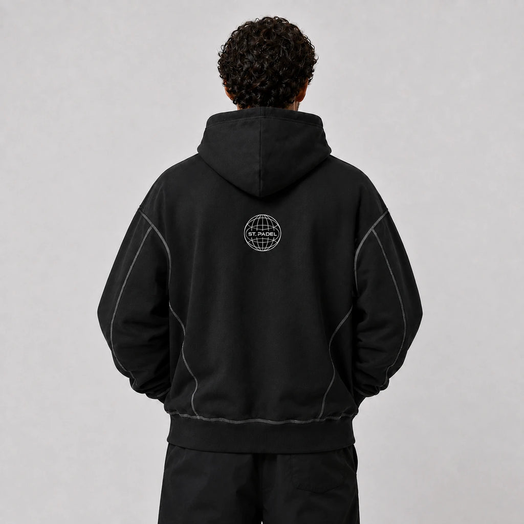 St. Padel Boxy Stich Hoodie. A person viewed from the back wearing a black pullover hoodie with a small circular white globe-style logo centered on the upper back. The hoodie has subtle light contrast stitching along the shoulders and sides, photographed against a plain light gray studio background.