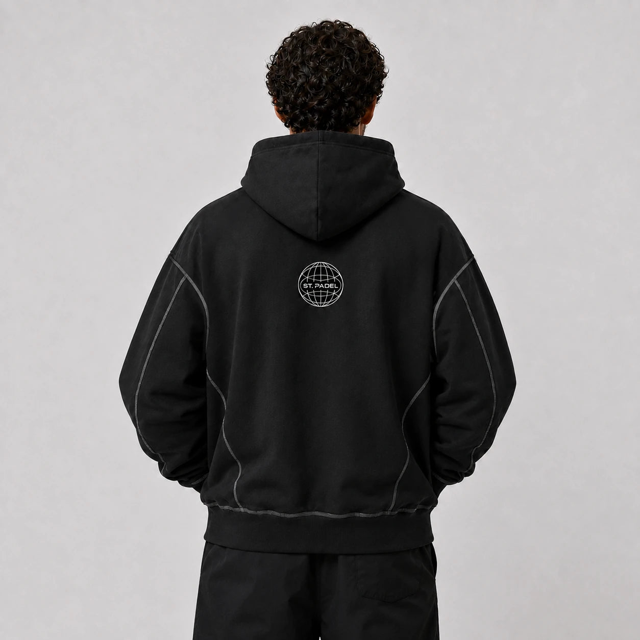 St. Padel Boxy Stich Hoodie. A person viewed from the back wearing a black pullover hoodie with a small circular white globe-style logo centered on the upper back. The hoodie has subtle light contrast stitching along the shoulders and sides, photographed against a plain light gray studio background.