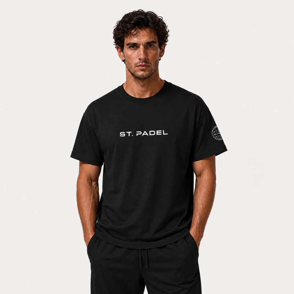 St. Padel Lifestyle Cotton T-Shirt. A man wearing a black short-sleeve t-shirt with 'ST. PADEL' printed in white on the chest and a circular logo on the left sleeve, paired with black pants, against a light off-white background.
