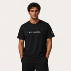 St. Padel Lifestyle Cotton T-Shirt. A man wearing a black short-sleeve t-shirt with 'ST. PADEL' printed in white on the chest and a circular logo on the left sleeve, paired with black pants, against a light off-white background.