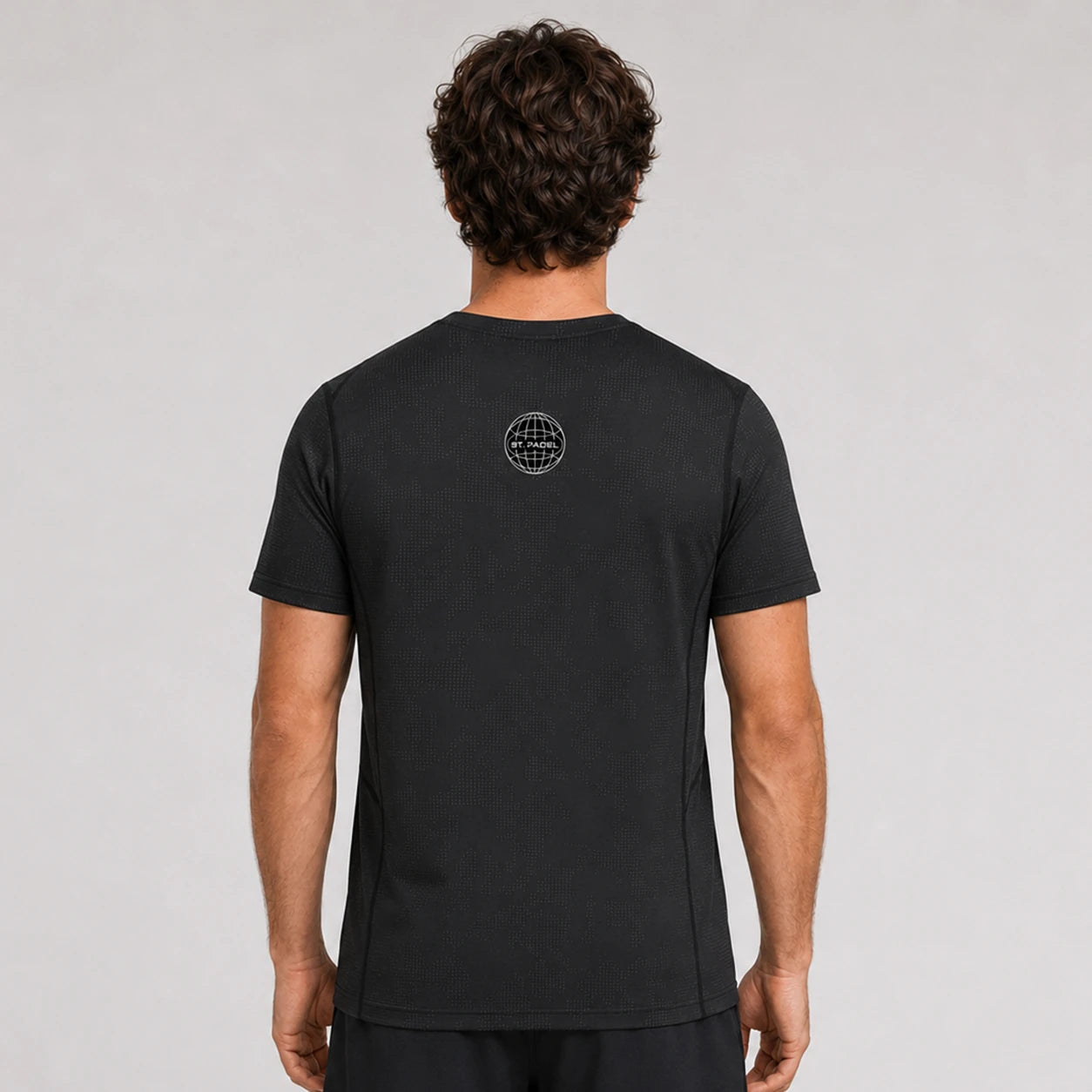 St. Padel Performance T-Shirt. A man viewed from behind wearing a black short-sleeve t-shirt with a small circular logo on the upper back, set against a plain light gray studio background.