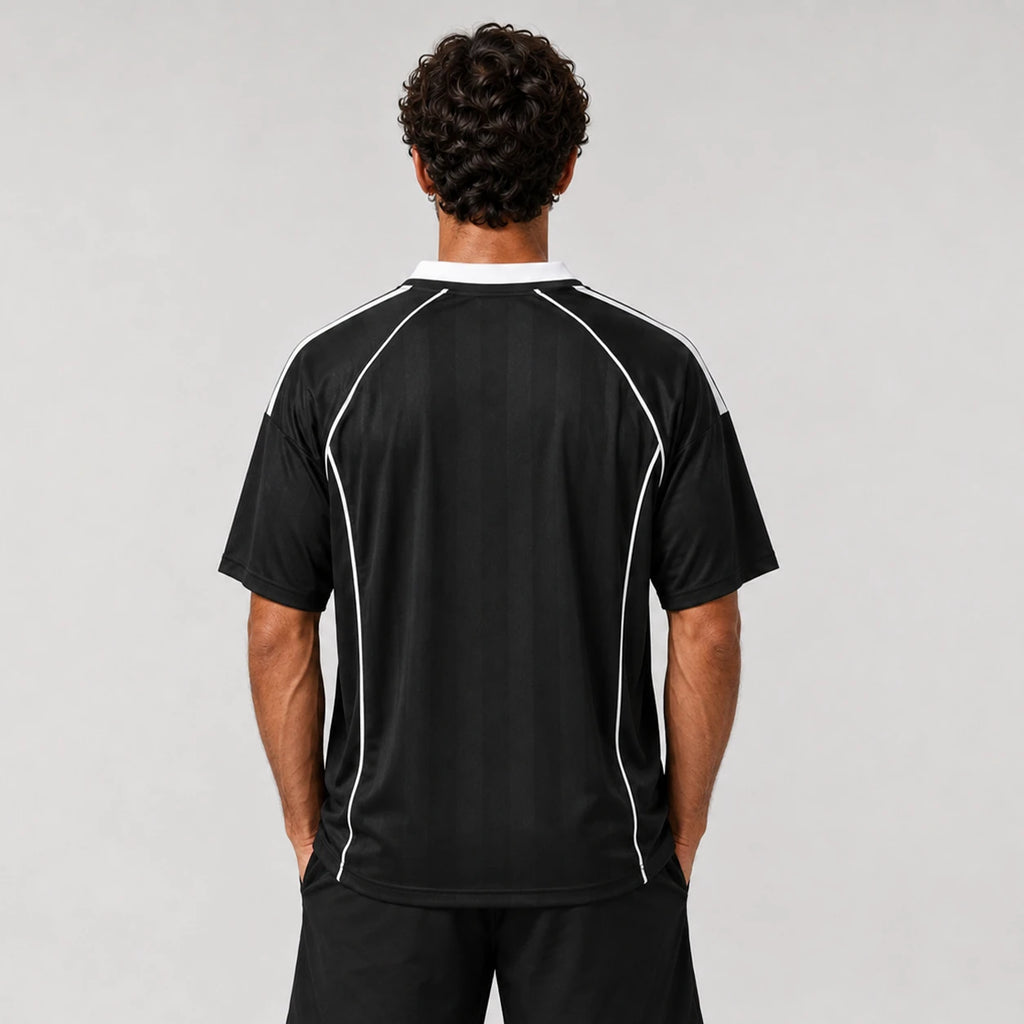 St Padel Shadow Stripe Jersey. A man viewed from behind wearing a black short-sleeve athletic jersey with white piping details along the shoulders and sides, paired with black shorts, against a light gray studio background.