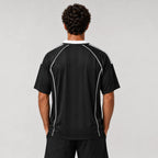 St Padel Shadow Stripe Jersey. A man viewed from behind wearing a black short-sleeve athletic jersey with white piping details along the shoulders and sides, paired with black shorts, against a light gray studio background.