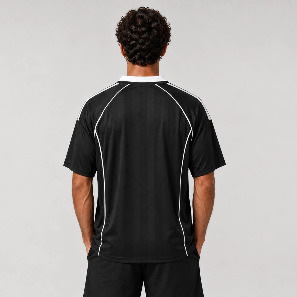St Padel Shadow Stripe Jersey. A man viewed from behind wearing a black short-sleeve athletic jersey with white piping details along the shoulders and sides, paired with black shorts, against a light gray studio background.