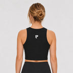 St. Padel Women's Crewneck Crop Tank Top. A woman viewed from the back wearing a black athletic crop top with a small white stylized logo on the upper back, paired with black bottoms, against a light gray studio background.