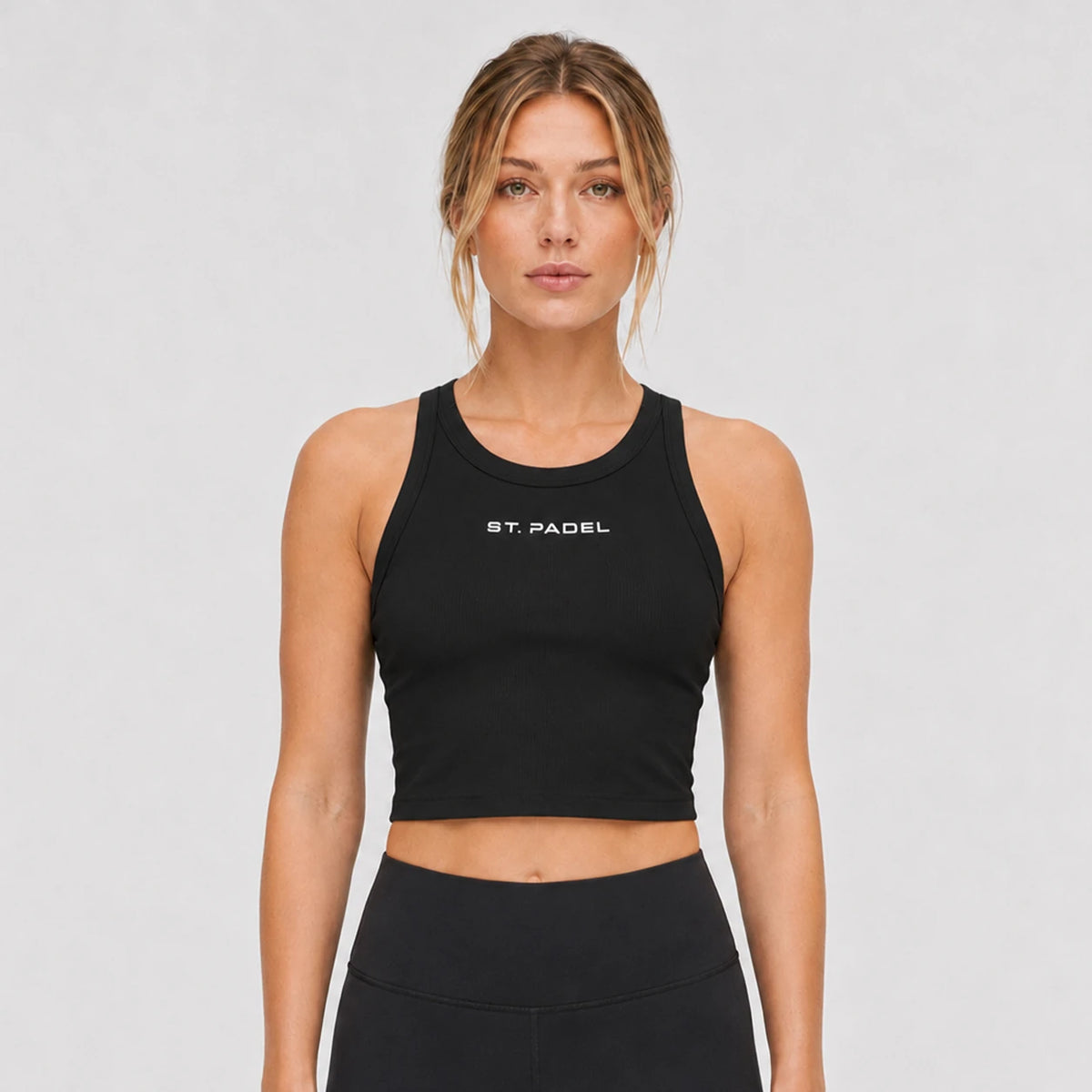 St. Padel Women's Crewneck Crop Tank Top. A woman modeling a black athletic crop top with 'ST. PADEL' printed in white on the chest, paired with black athletic bottoms, against a light gray studio background.