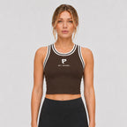 St Padel Women's Performance Ringer Tank. A woman modeling a dark brown athletic crop top with white trim and a white St. Padel logo on the chest, paired with black athletic leggings, against a light gray studio background.